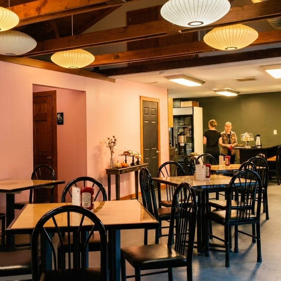 A restaurant dining room with pink and green walls, wooden tables, black chairs, and two women at a counter.