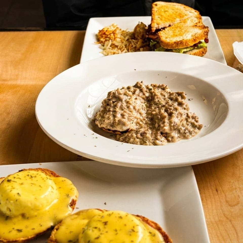 Three breakfast plates on a wooden table: eggs Benedict, biscuits and gravy, and a sandwich with hash browns.