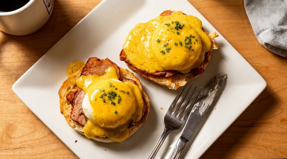 Two eggs Benedict with hollandaise sauce and chives on a white plate with a fork and knife.